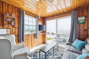 A wooden interior with a TV, a sofa, a chair, and a view of the beach.