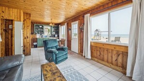 A cabin with a wooden interior, a kitchen area, a seating area with a sofa and armchair, and a large window overlooking a beach.