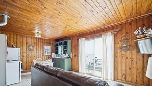 En stuga med träinredning, ett litet kök, en sittgrupp med soffa och utsikt över stranden genom ett stort fönster.