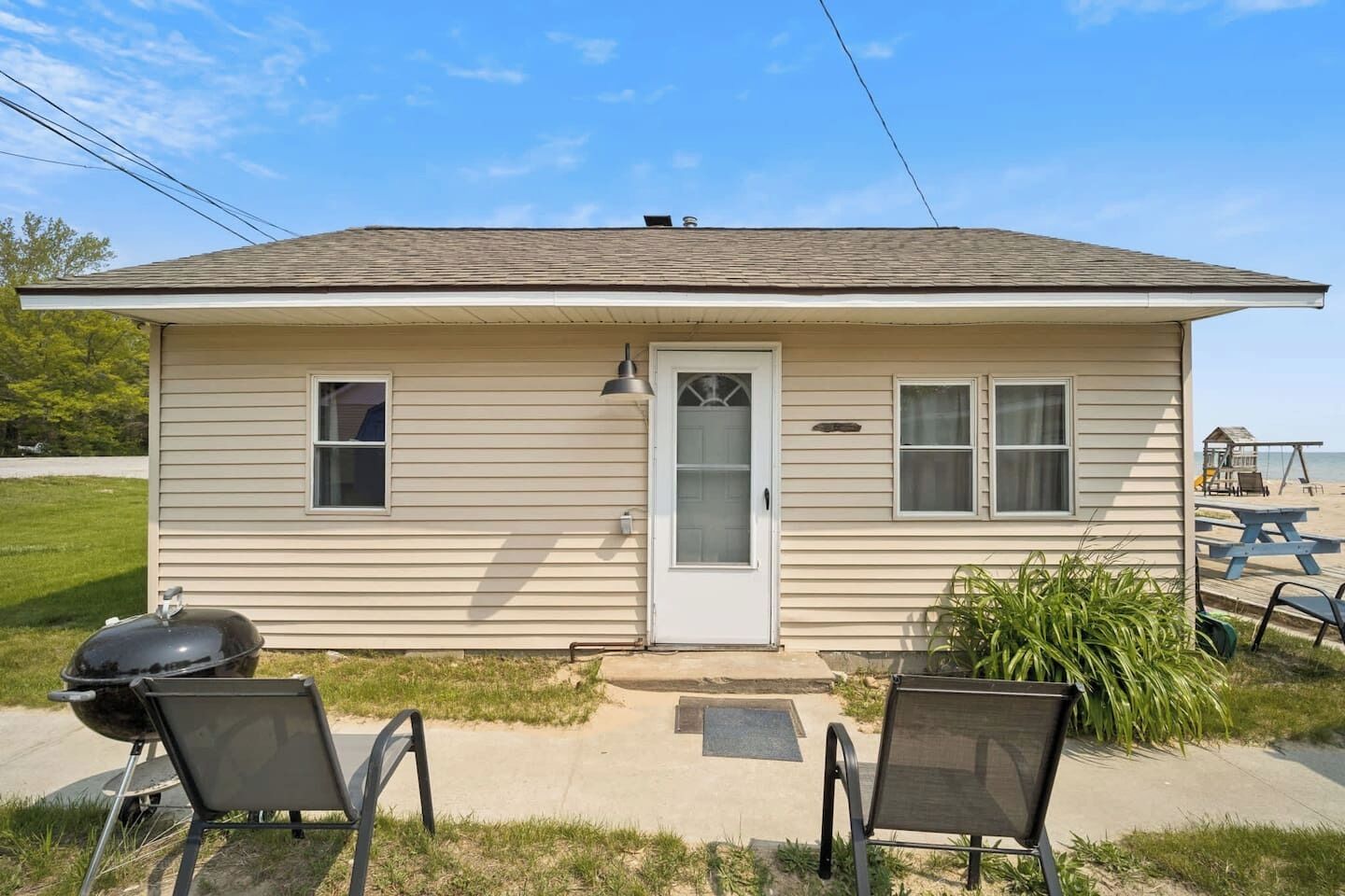 A single-story house with a front door, two windows, a small porch, and a barbecue grill.