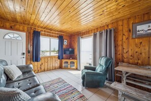 Een cabin interieur met een houten plafond, een leren bank, een smaragdgroene fauteuil, een televisie op een houten kast, en uitzicht door een groot raam.