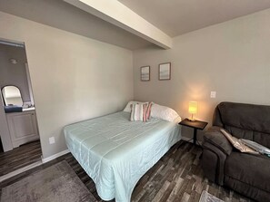 A bedroom with a bed, a nightstand, a lamp, a couch, and a small bathroom.