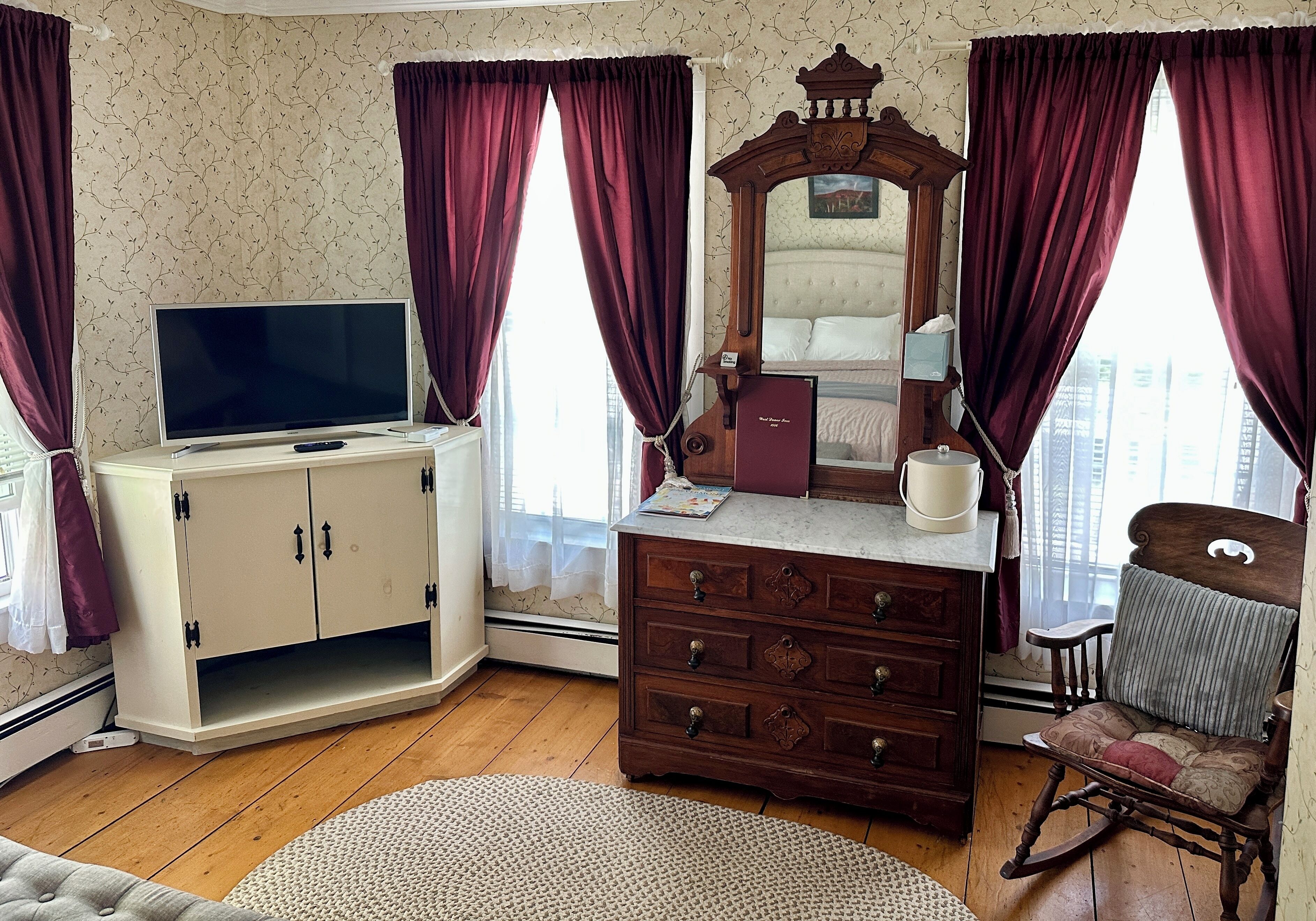 A room with a television, a dresser, a mirror, a rocking chair, and a window with curtains.