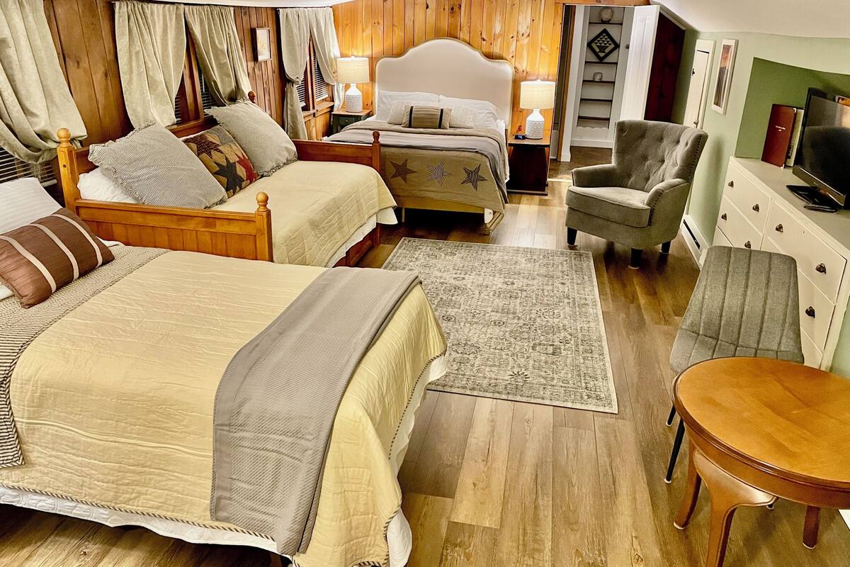 A hotel room with two beds, a sitting area with a chair and a small table, a television, and a bookshelf.