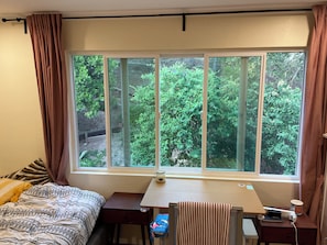 A bedroom with a large window, a bed with patterned bedding, a desk with a chair, and a small table with a lamp.