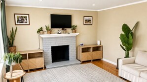 A living room with a fireplace, a flat-screen TV mounted above it, and a sofa. There are potted plants and a small table with a clock. - Spacious 4-bedroom house with AC in tranquil Orinda (Orinda)