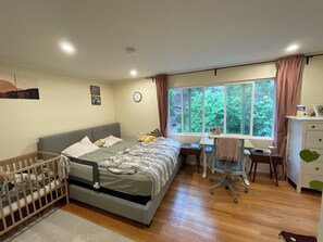 A bedroom with a bed, a desk, a chair, a crib, a window with curtains, and a clock on the wall.
