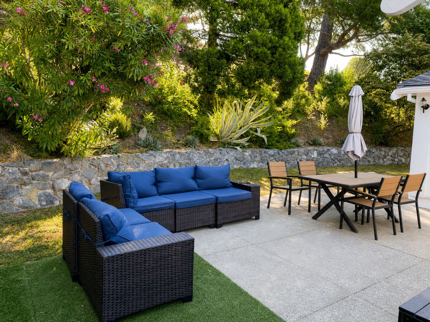 An outdoor patio area with a stone wall, a wicker sofa with blue cushions, a wooden table with chairs, and a white umbrella.