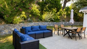 An outdoor patio area with a stone wall, a wicker sofa with blue cushions, a wooden table with chairs, and a white umbrella.