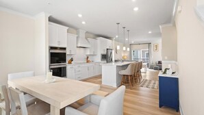 Une cuisine moderne avec des meubles blancs, un coin repas doté d’une table et de chaises aux couleurs claires, et un petit coin bar avec des tabourets.