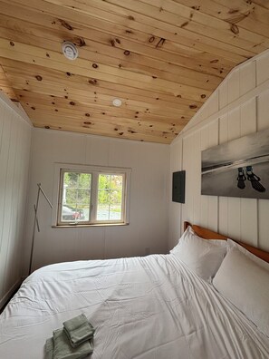 Quarto com teto de madeira, janela, cama com roupa de cama branca e luminária fixada na parede.
