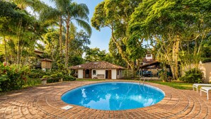 Una piscina de ladrillo circular rodeada de abundante vegetación y una pequeña construcción tipo cabaña.