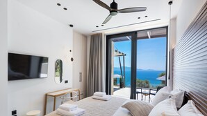 A modern hotel room with a bed, a TV, a desk, and a balcony with a view of the sea.