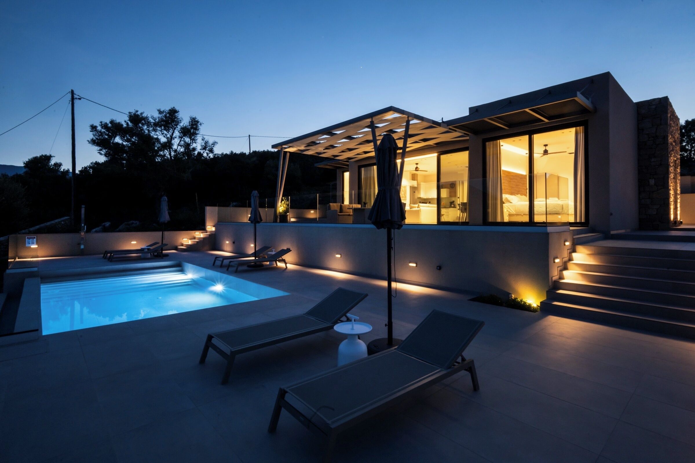 Una casa moderna con piscina, mobiliario exterior e interiores bien iluminados.