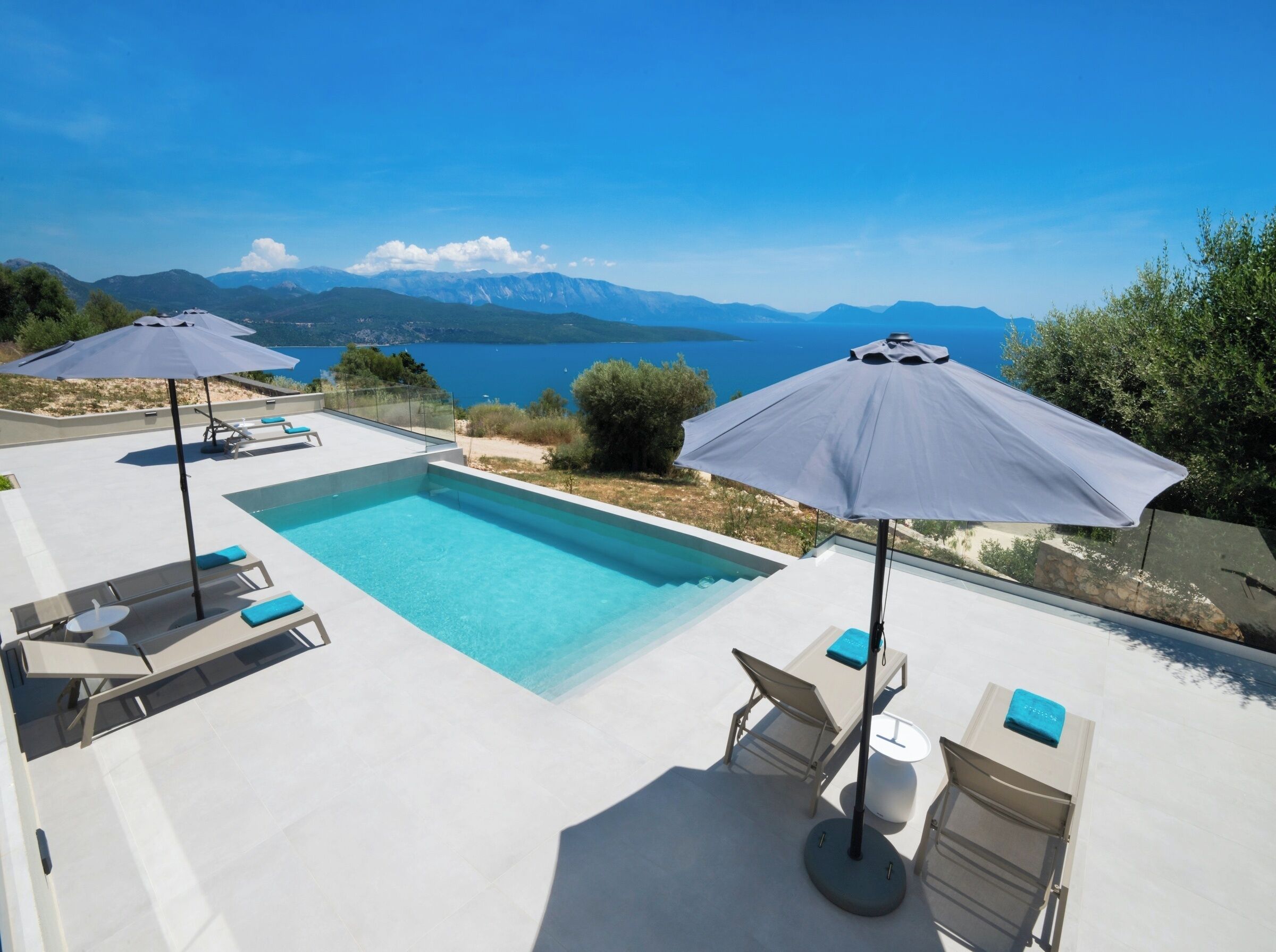 Zona de piscina con sillones reclinables y sombrillas, con vistas panorámicas a montañas y una masa de agua.