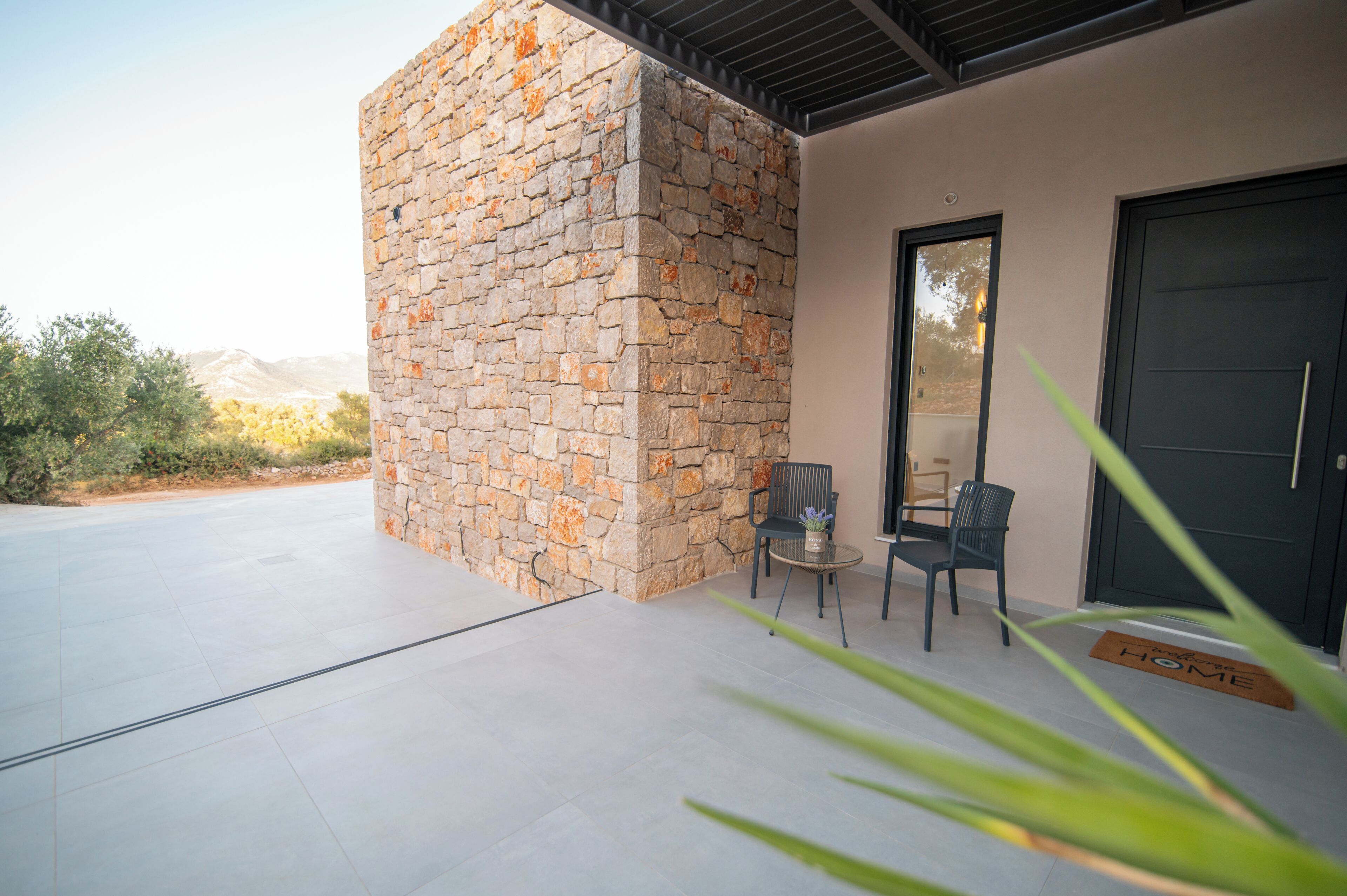 A modern building with a stone facade, a door, and a patio area with chairs and a doormat.