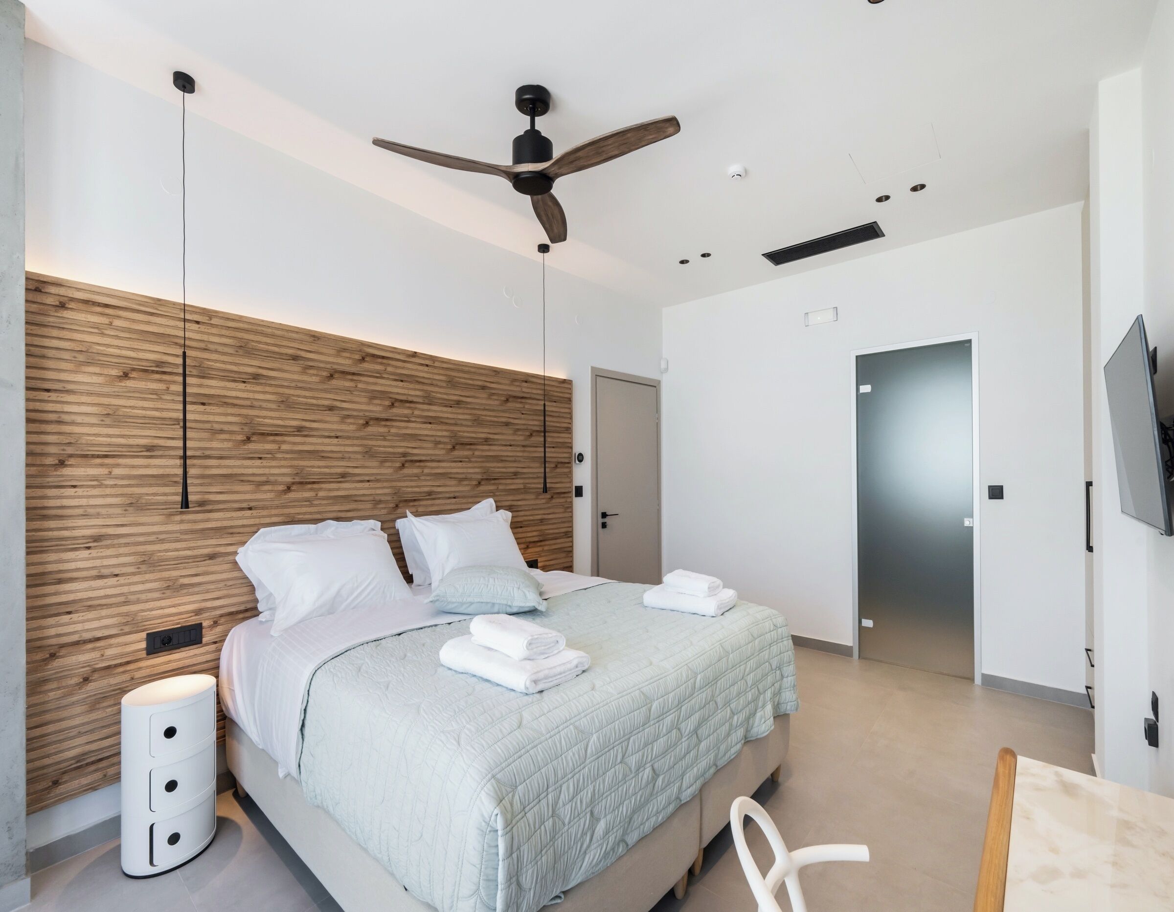 Une chambre moderne avec une tête de lit en bois, un lit avec une literie blanche, un ventilateur de plafond et un téléviseur fixé au mur.