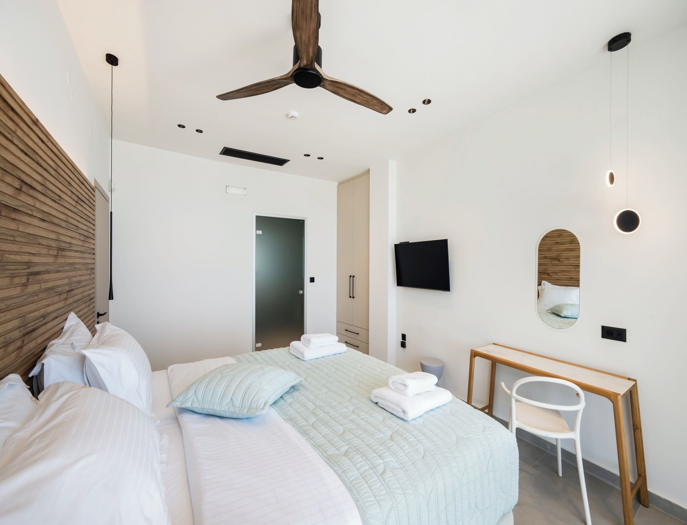 Een moderne slaapkamer met een bed, nachtkastje, spiegel, plafondventilator en een aan de muur bevestigde televisie.