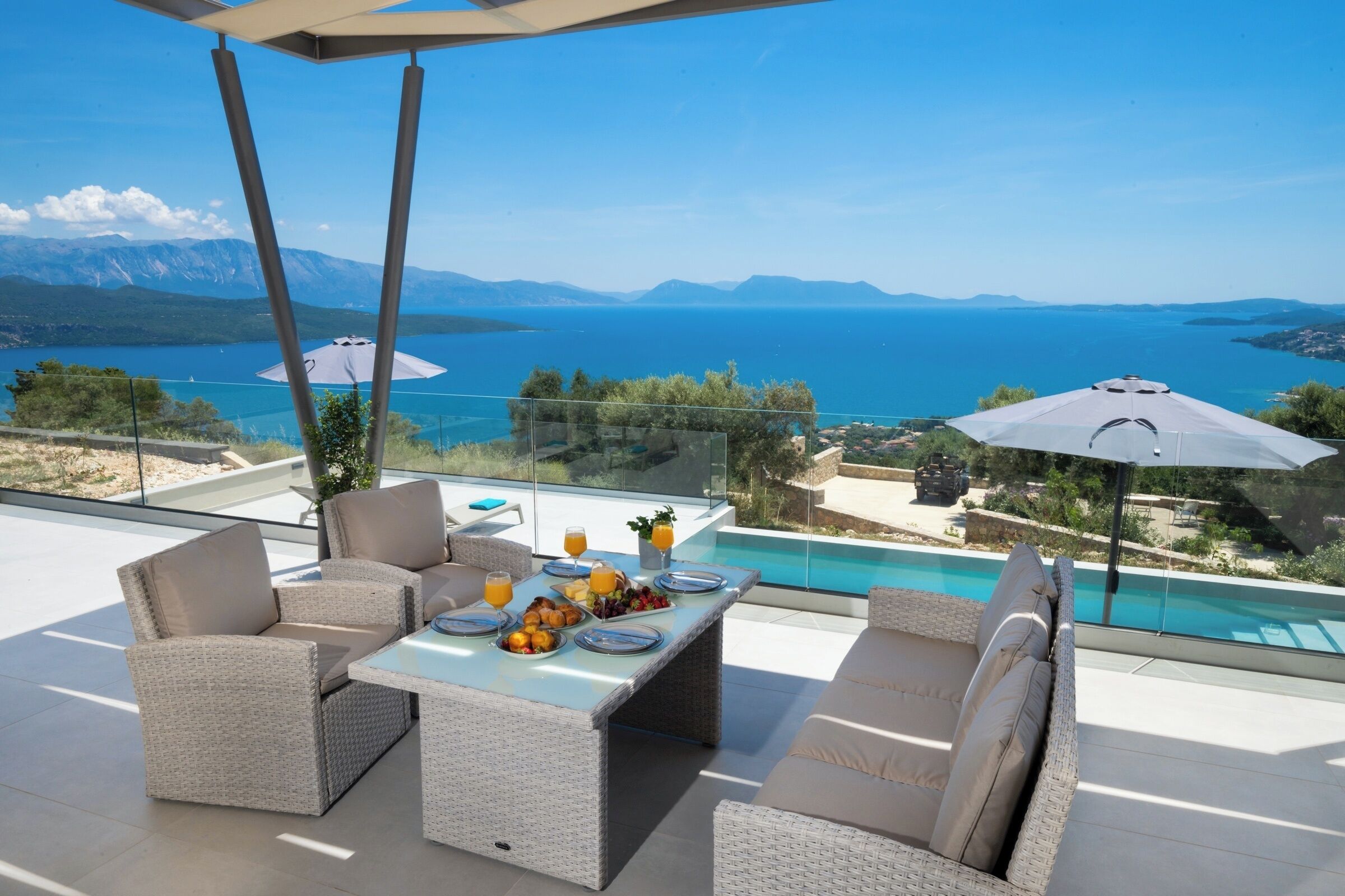 A terrace with wicker furniture, a dining table set with plates and drinks, and a view of a body of water and mountains.