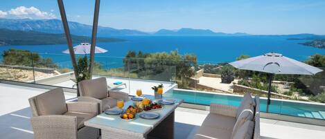 A terrace with wicker furniture, a dining table set with plates and drinks, and a view of a body of water and mountains.