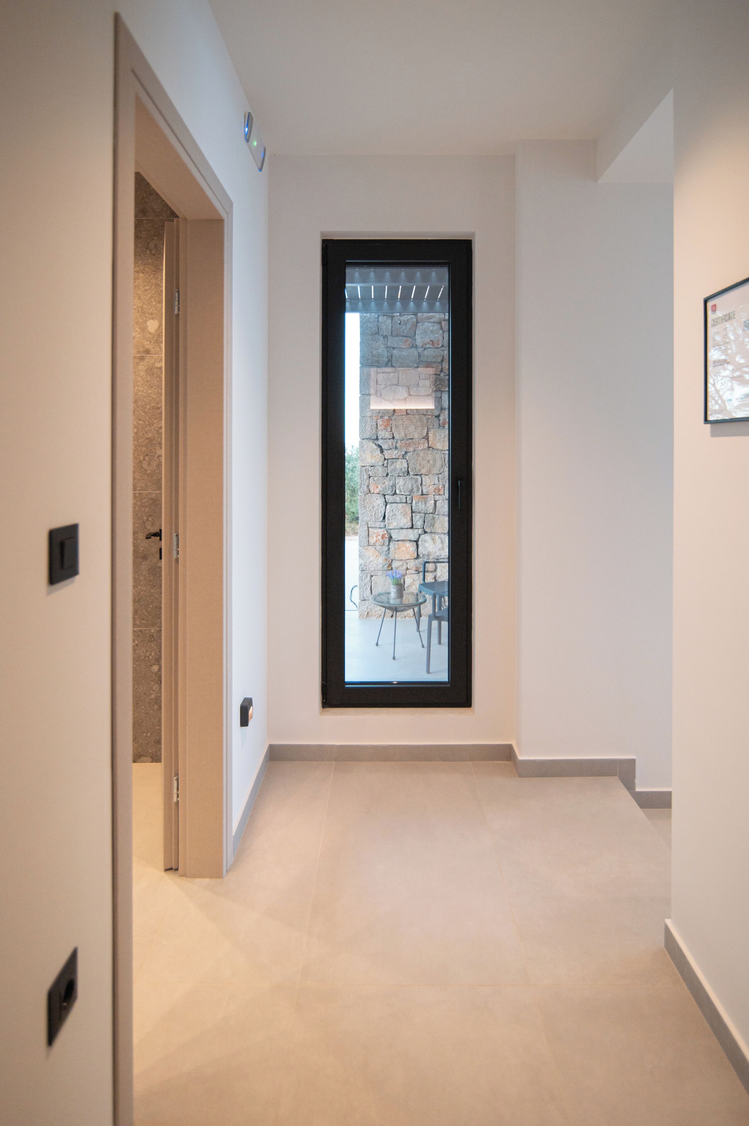 A hallway with a stone wall and a window, leading to an outdoor area with chairs.