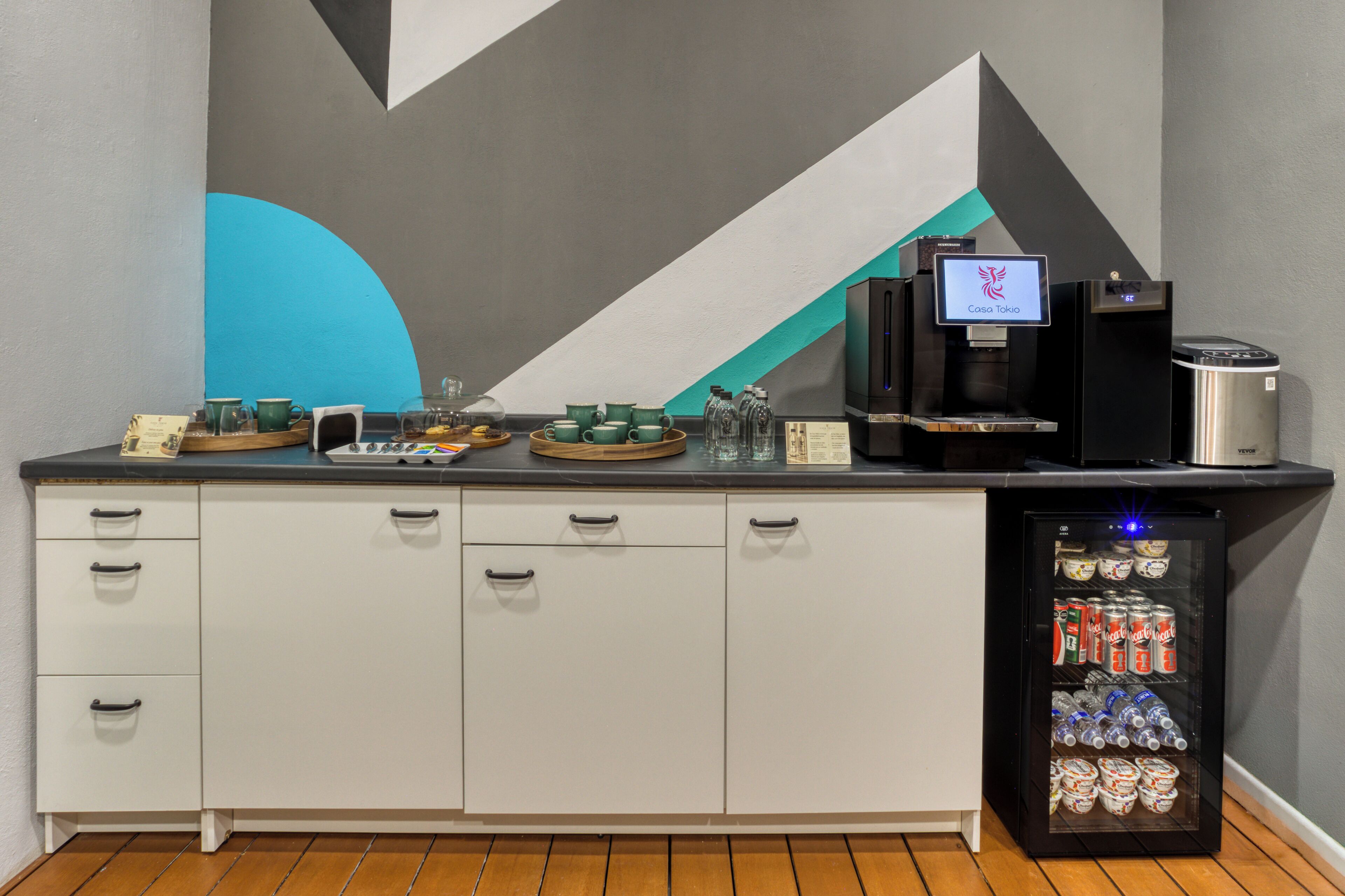 A kitchenette with a coffee machine, a refrigerator stocked with beverages, and a wall with a geometric design.
