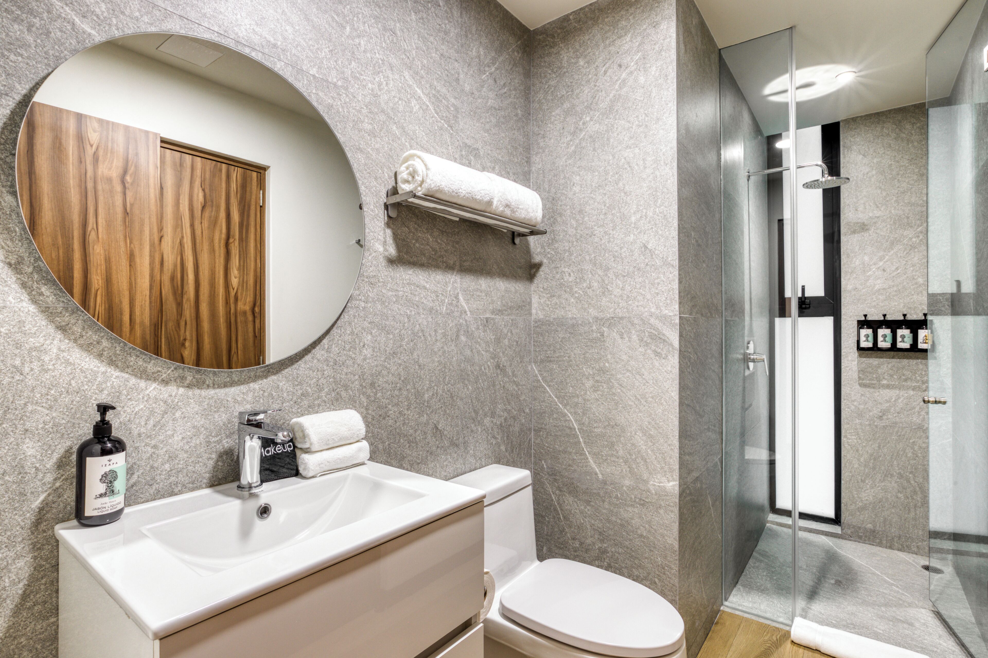 Un baño moderno con un espejo redondo, un mueble de madera, un lavabo blanco, un inodoro y una ducha con mampara de vidrio.