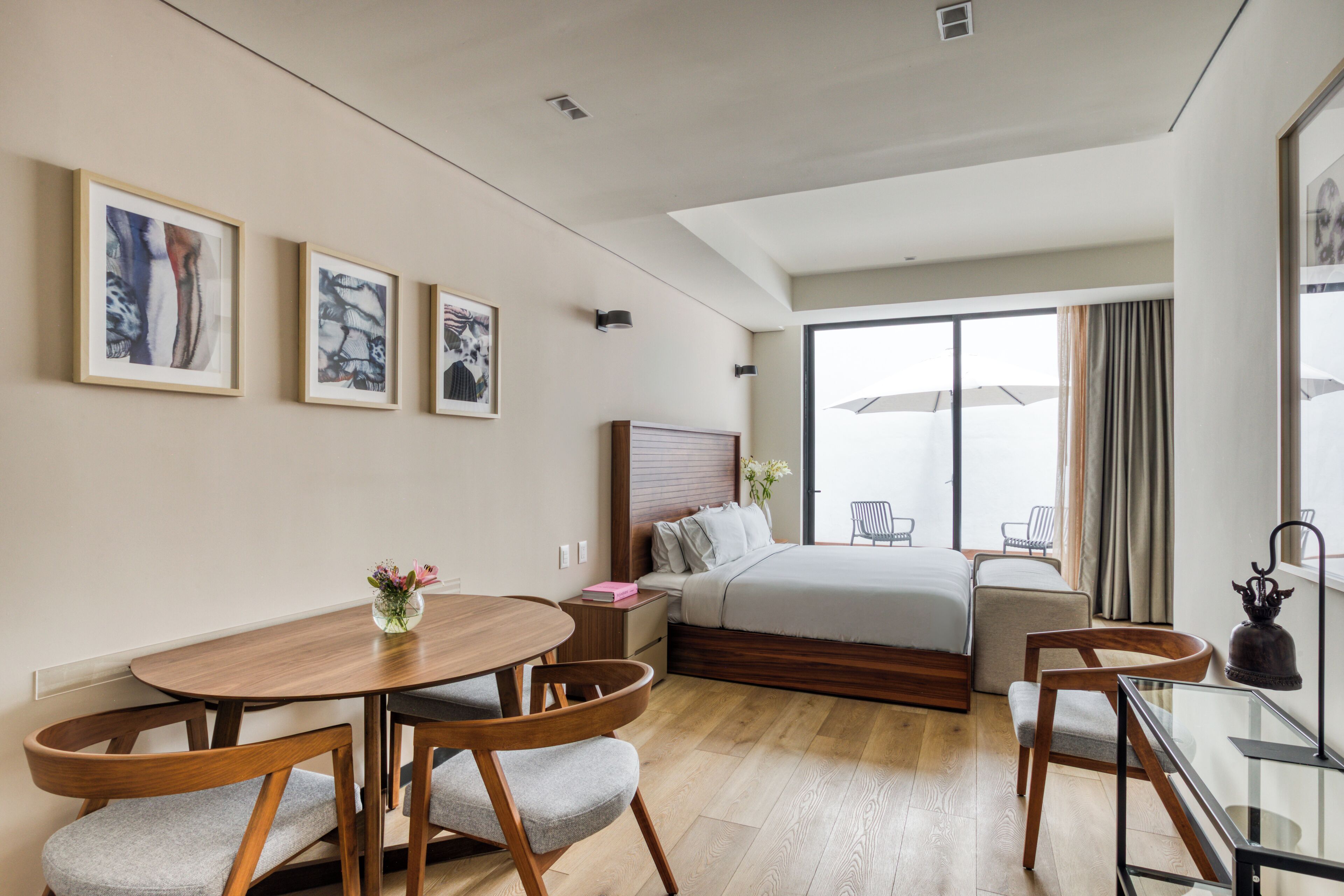 A hotel room with a bed, a desk, a chair, a dining table, and artwork on the wall.