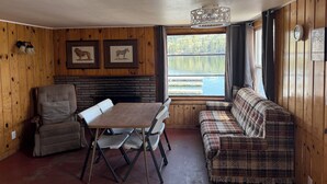 A cabin interior with a dining table, two chairs, a sofa, wooden walls, a fireplace, framed animal pictures, and a window with a view of a lake.