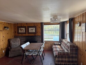 Interno di una baita con tavolo da pranzo, due sedie, divano, pareti in legno, camino, quadri con immagini di animali e finestra vista lago.
