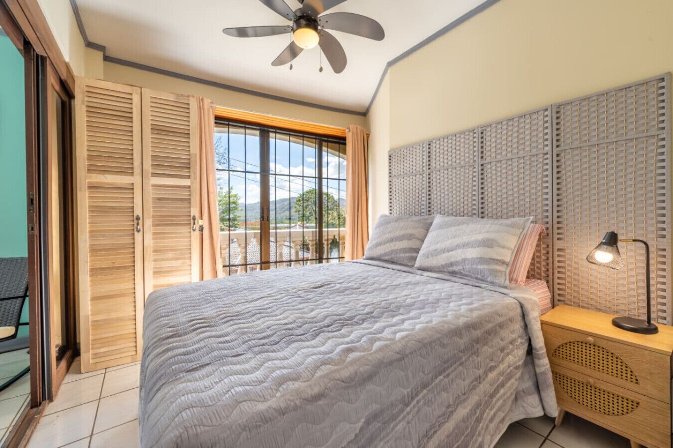 Une chambre à coucher comprenant un lit, une table de chevet, une lampe, un ventilateur de plafond, une fenêtre avec des rideaux et une porte coulissante.
