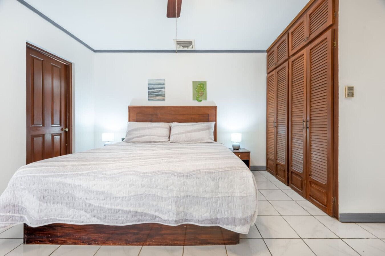 A bedroom with a wooden bed, two bedside lamps, a wooden wardrobe, and a ceiling fan.