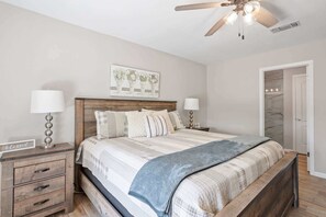 Un dormitorio con una cama de madera, dos mesitas de noche con lámparas, un ventilador de techo, una puerta y un cuadro en la pared.