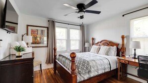 Ein Schlafzimmer mit einem hölzernen Bett, einem Schreibtisch, einem Stuhl, einem Deckenventilator, einem Fernseher und einem Fenster mit Vorhängen.