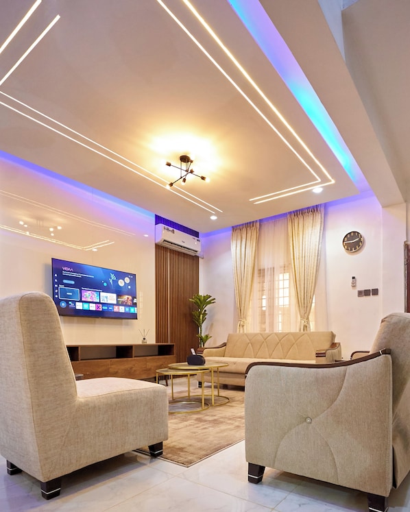 Ein modernes Wohnzimmer mit Flachbildfernseher, zwei Sesseln, einer Couch, einem Couchtisch und einer Deckenleuchte.