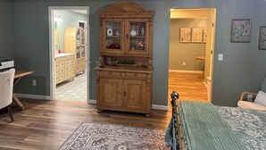 Une chambre avec une commode en bois, un lit recouvert d’une couverture verte, un plancher en bois et un couloir menant à une autre pièce.