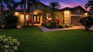 A residential house with a well-maintained lawn, palm trees, and a driveway leading to the garage.