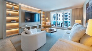 Ein modernes Hotelzimmer mit Flachbildfernseher, einer Couch, einem Sessel, einem Couchtisch und einem Bücherregal.