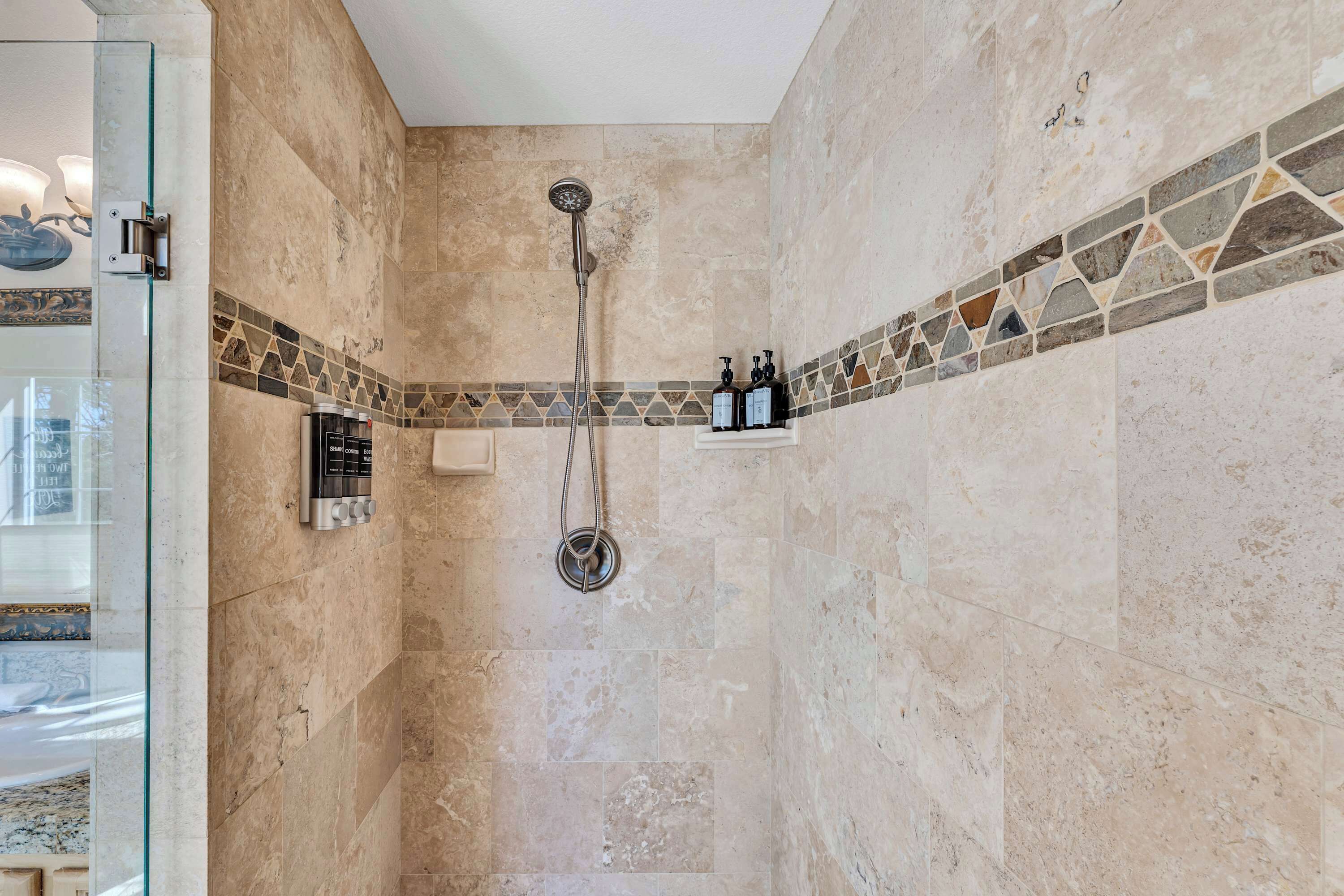 A shower with a handheld showerhead and a wall-mounted faucet, a glass shower door, and a tiled shower area with a decorative border.