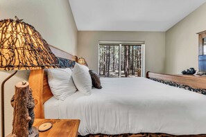Ein Schlafzimmer mit einem großen Bett, einem hölzernen Kopfteil, einem Nachttisch, einer Lampe und Blick auf Bäume durch ein Fenster.
