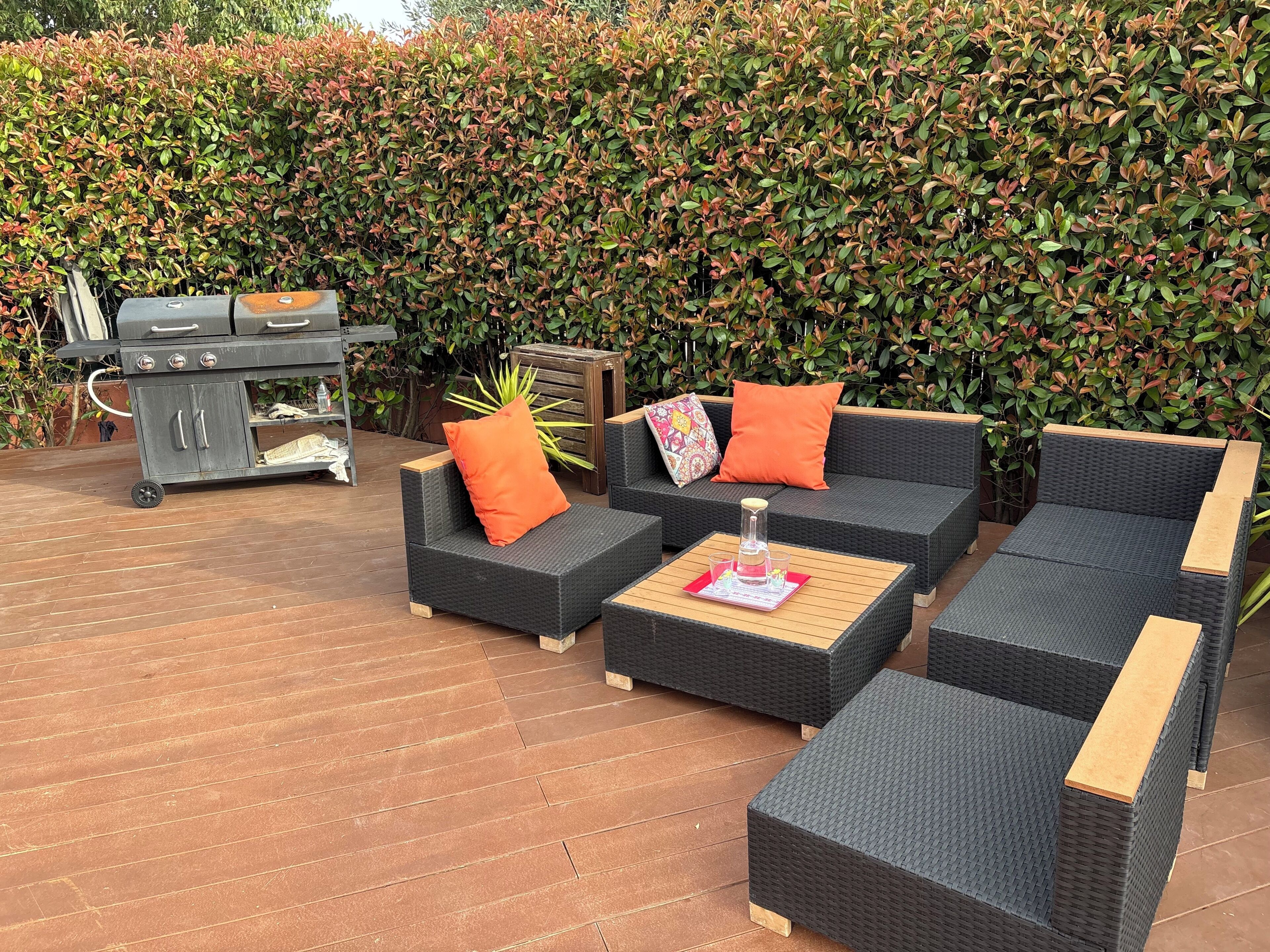 An outdoor patio with wicker furniture, a grill, and a hedge in the background.