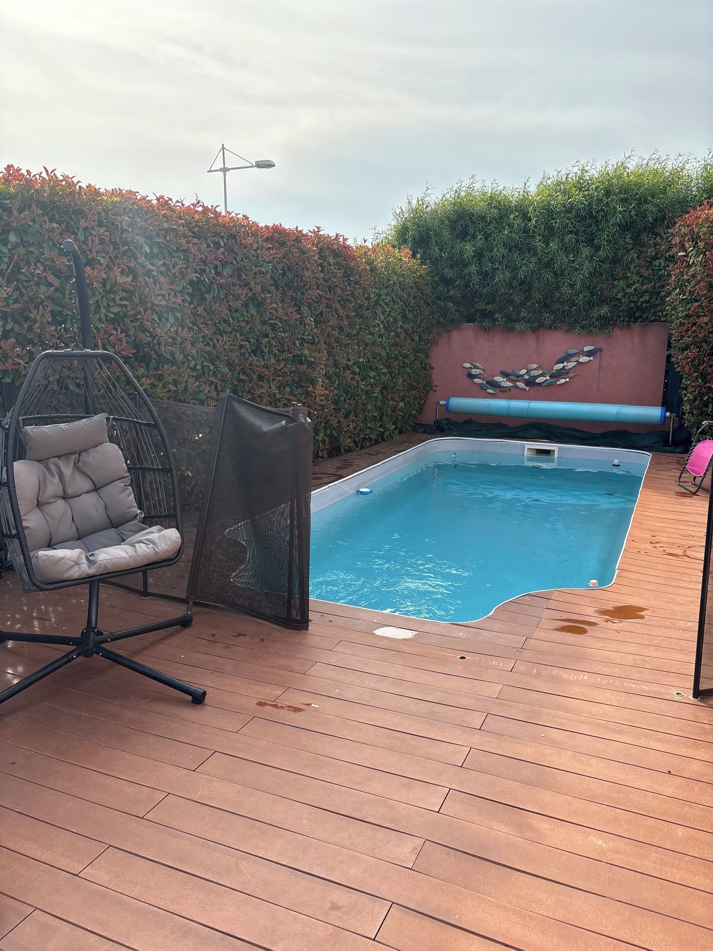 A swimming pool with a wooden deck, a hanging chair, and a wall with a decorative stone design.