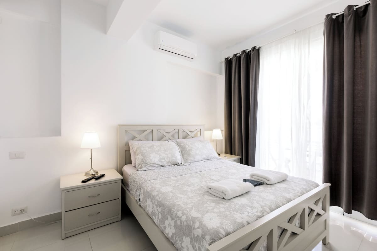 Quarto com cama, mesas de cabeceira, ar condicionado e janela com cortinas.