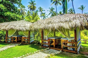 Duas cabanas com telhados de palha, bancos de madeira e cortinas, cercadas por vegetação exuberante e palmeiras.