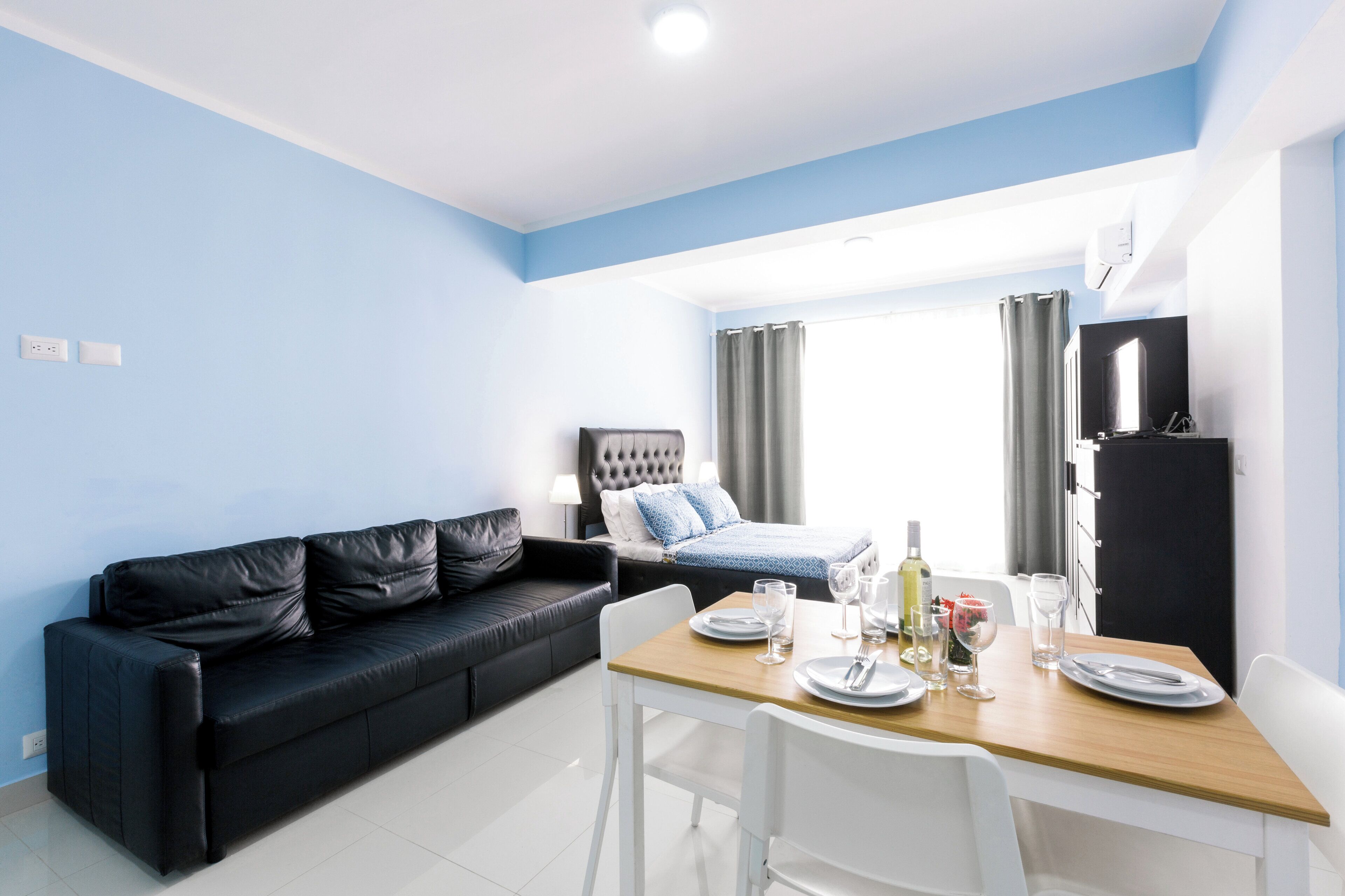A modern hotel room with a black leather sofa, a wooden dining table set for two, a bed with a headboard, and a black wardrobe.
