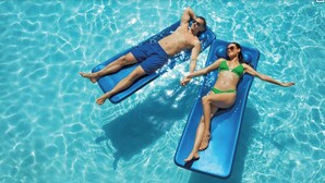 Duas pessoas relaxando em espreguiçadeiras azuis flutuantes em uma piscina azul cristalina.