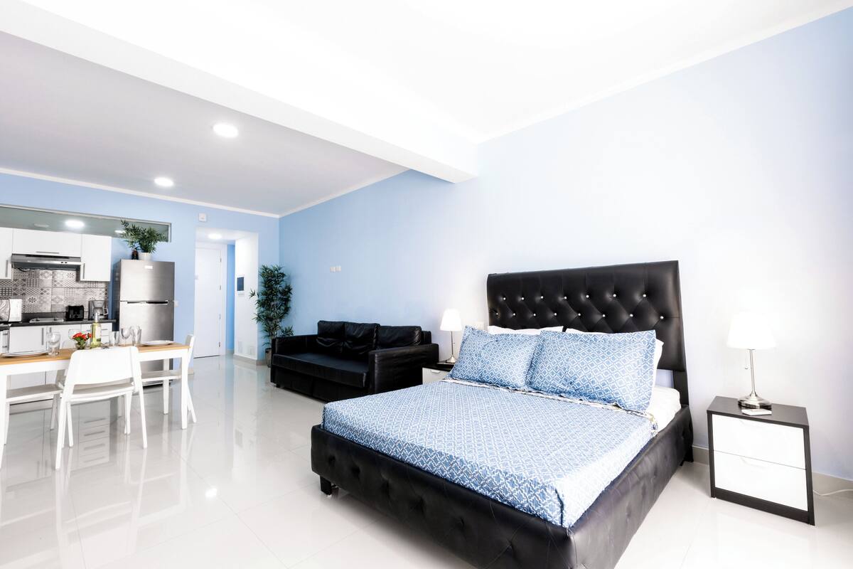 Quarto moderno com cama grande, sofá independente, mesa de jantar com cadeiras e cozinha compacta ao fundo.