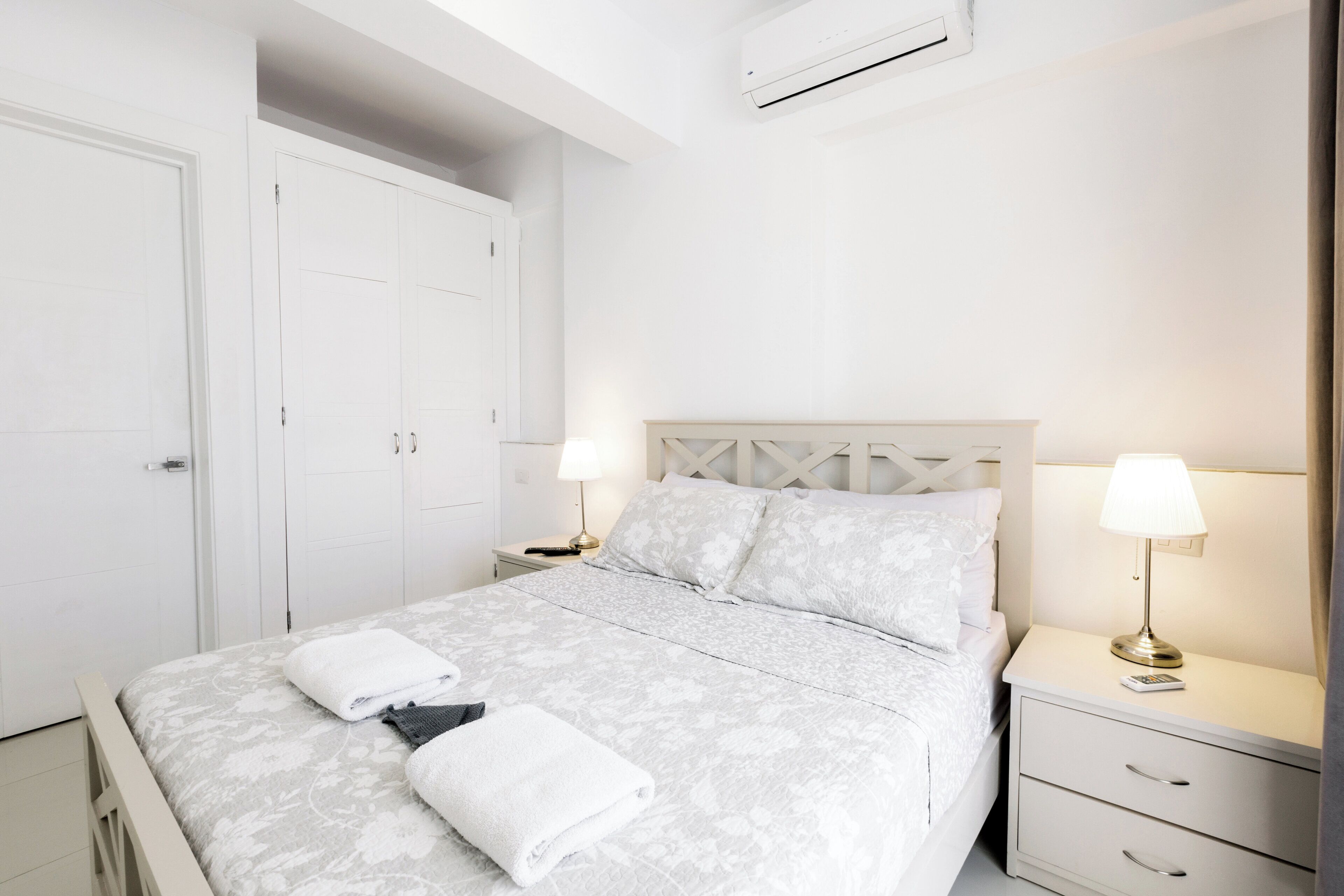 A bedroom with a bed, two bedside tables, a nightstand, a wardrobe, and an air conditioning unit.