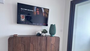 A modern living room with a flat-screen TV mounted on the wall, a wooden console table with decorative items, and a large window. - The View 108 - Stylish apartment near the beach in Baja Sardinia (Baja Sardinia)