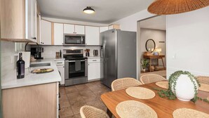 A modern kitchen with white cabinets, a stainless steel refrigerator, a microwave, and an oven. There is a dining area with a wooden table and woven chairs. - Hot Tub, Sauna & Game Room Mountain-View Home (Westminster)